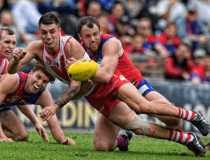Two AFL players low to the ground, one trying to grab the ball, the other tackling them to the ground