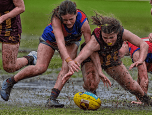 Two players in the WAWFL scramble in a tackle for the ball as mud and water splash around them during wet conditions